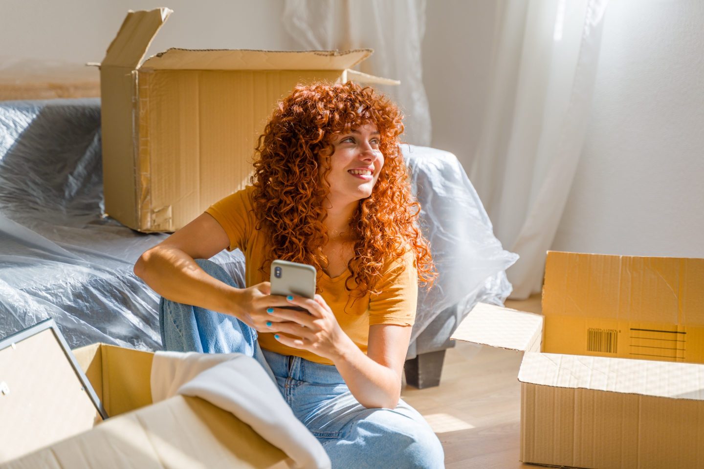 Young woman is using smartphone and smiling while taking a break from unpacking cardboard boxes in her new apartment