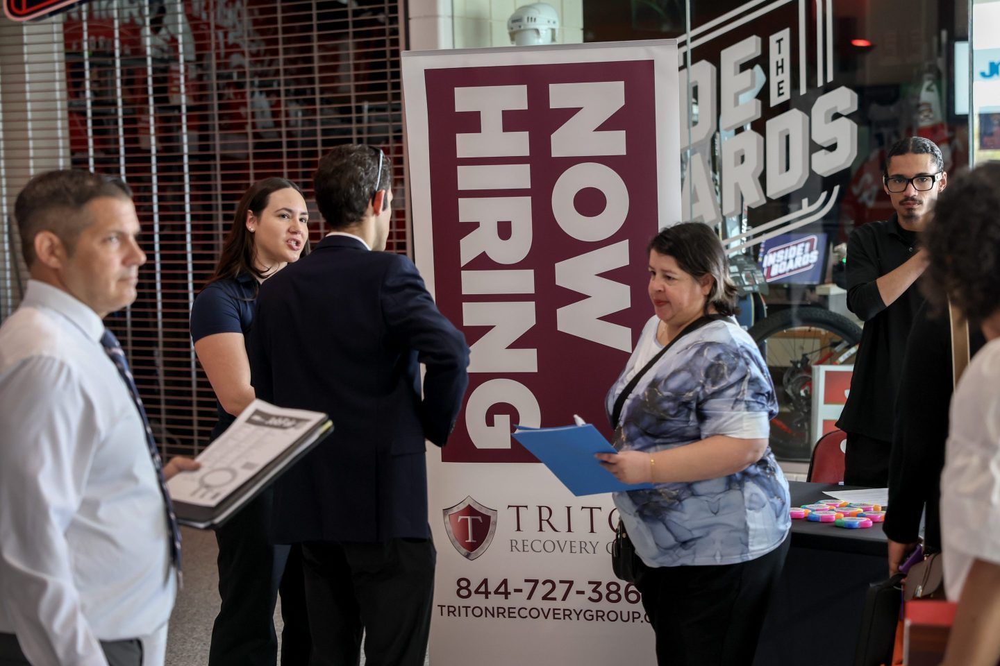 A picture of people milling about and talking with recruiters at a job fair with a "Now Hiring" sign in the background.