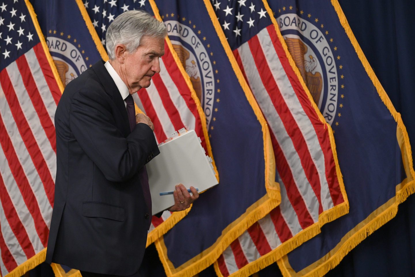 Federal Reserve Chair Jerome Powell walks to his left past several staggered American flags and flags with the emblem of the Federal Reserve.