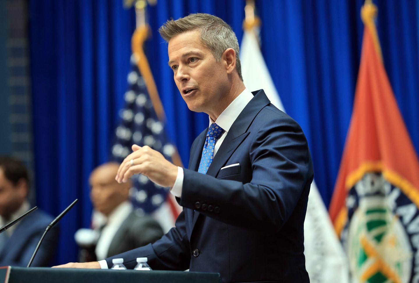 Sean Duffy speaks into a microphone on a podium in front of several flags on stage.
