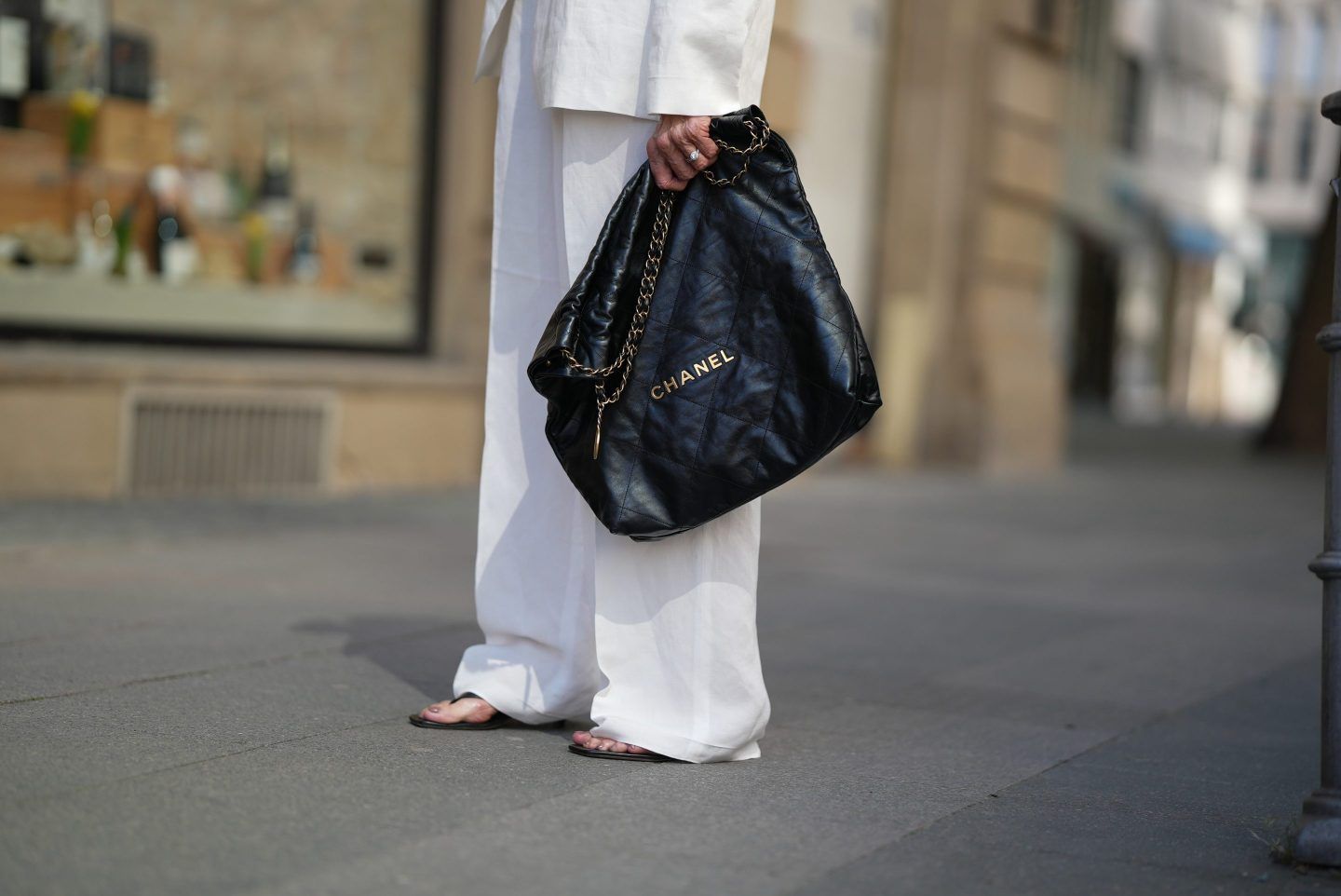Angelika Hanel is seen wearing a white linen suit blazer with lapel collar and two flap pockets from Massimo Dutti, a white linen pleated suit trousers from Massimo Dutti, black leather flip flop kitten heels from Toteme, black sunglasses with white trim and logo detail from Chanel, a black Chanel 22 handbag