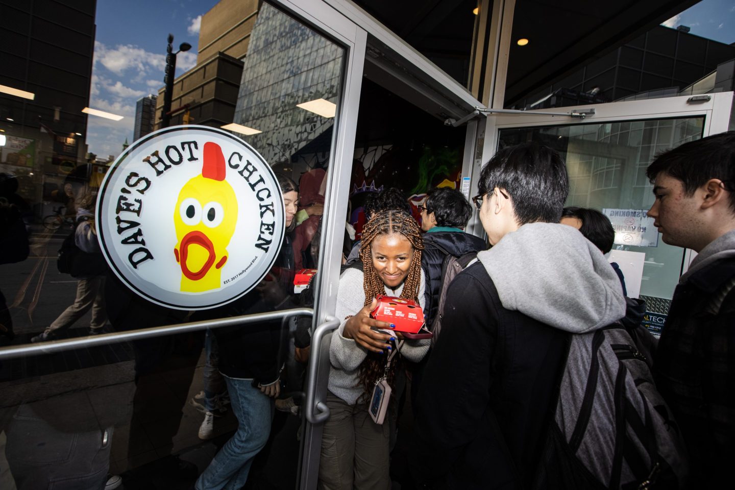 A woman squeezes through a door with her chicken sandwich as she exits a Dave's Hot Chicken with a line going out the door.