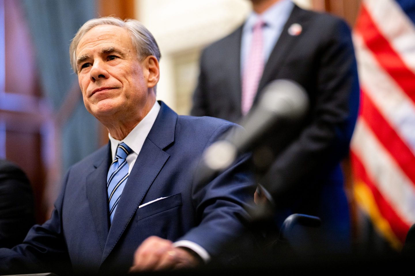 Greg Abbott, wearing a dark suit, sits behind a microphone and looks to his right.
