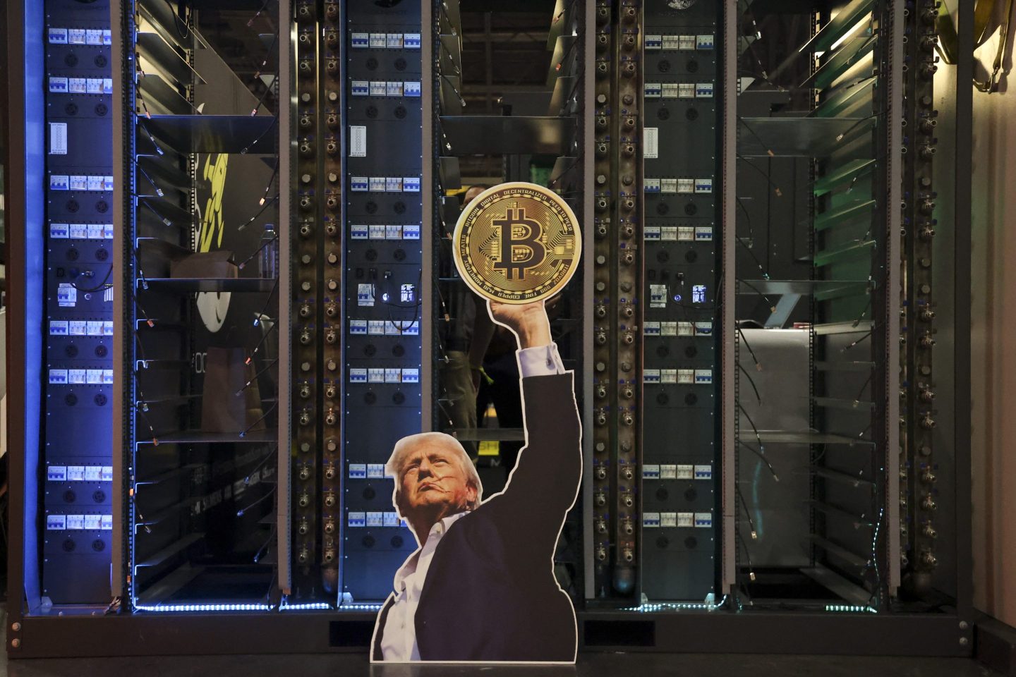 A cut-out of Trump holds up a bitcoin in front of some data servers