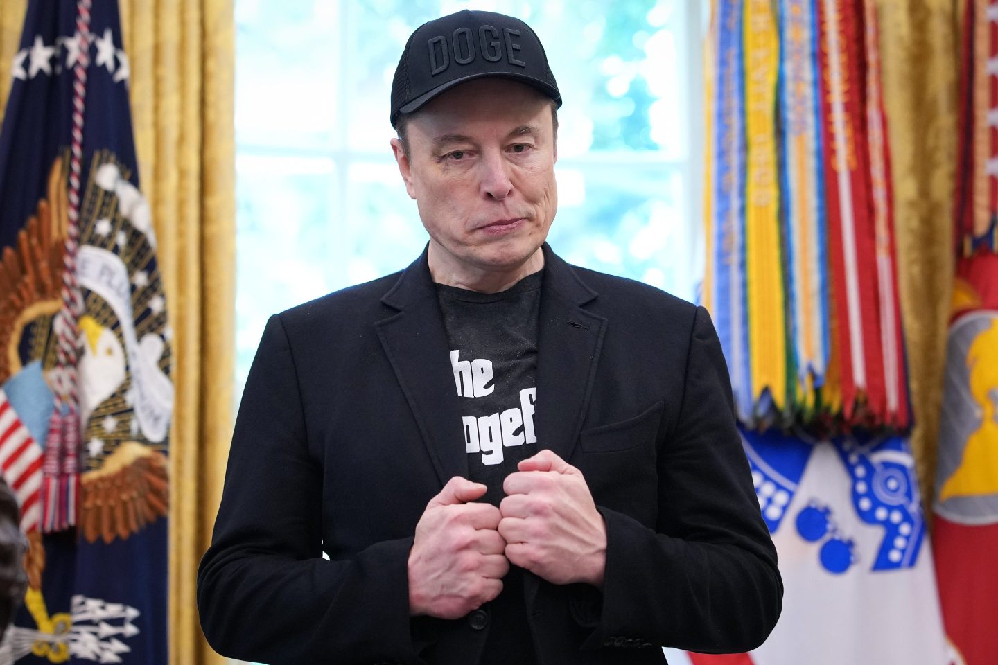 Elon Musk, wearing a DOGE hat and standing in the Oval Office, looks down and chews his lip.