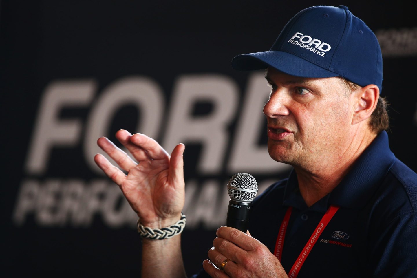 Jim Farley, wearing a blue "Ford" hat, speaks into a microphone and gestures with his hand.