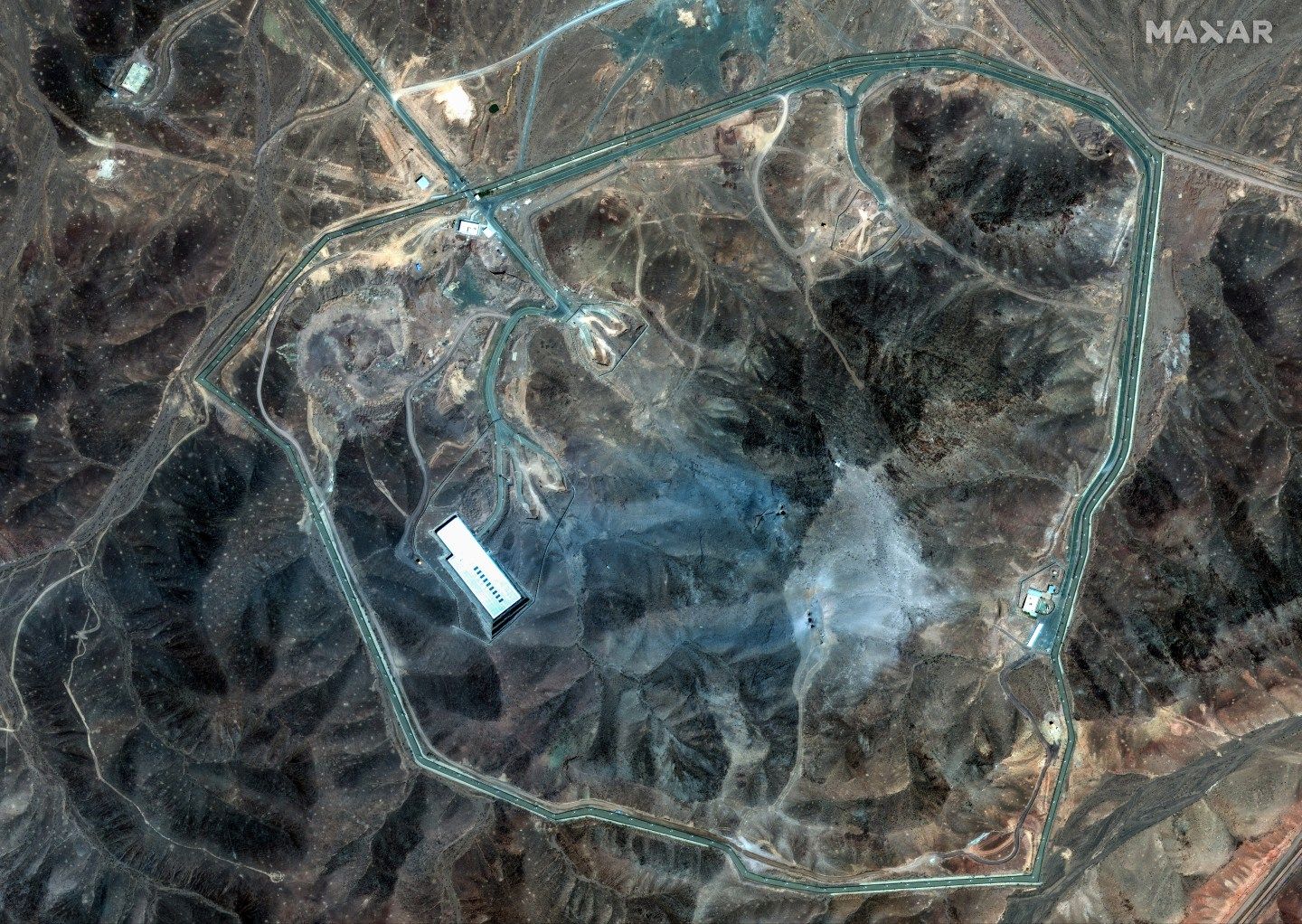 Satellite Imagery shows extensive damage at the Fordow underground complex in Iran. Several large craters are visible across the ridge, and a wide area is covered in grey-blue ash, consistent with airstrike aftermath. Satellite image (c) 2025 Maxar Technologies.