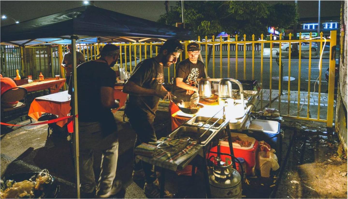 Men work in front of fryers and other cooking equipment under a tent at night.