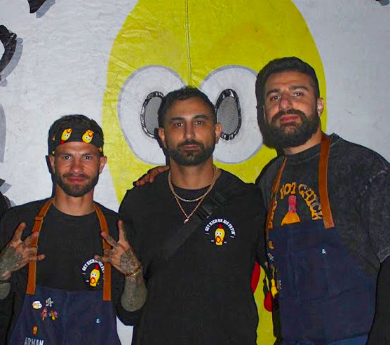 Three of the Dave's Hot Chicken founders posing in branded shirts and aprons in front of a banner with the company's mascot, a yellow rubber chicken.