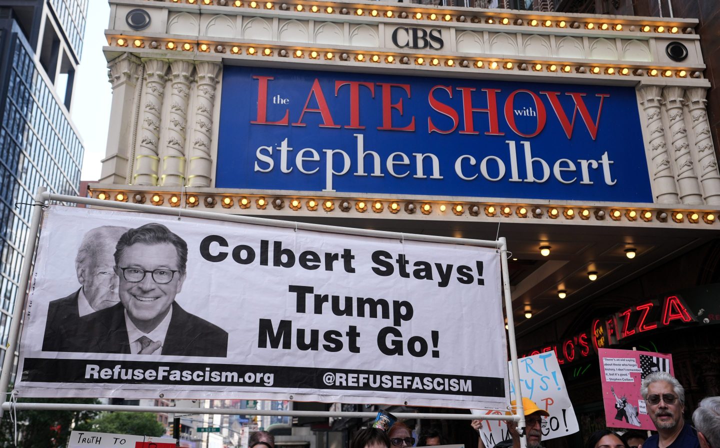 Someone holds a sign that says "Colbert Stays! Trump Must Go!" outside the Ed Sullivan Theater