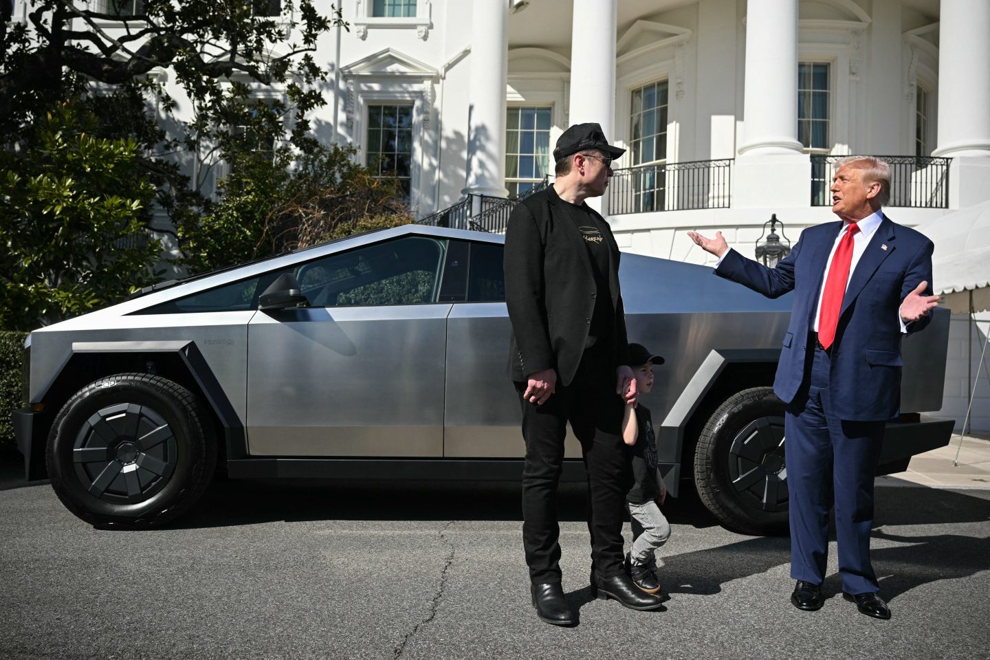 Elon Musk and Donald Trump look at each other while standing in front of a Cybertruck in the White House driveway.