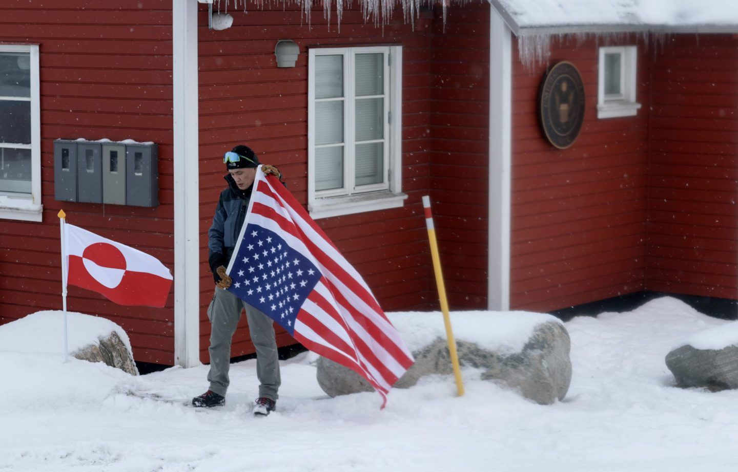 A greenlander on a snowy day hoists the American flag upside down next to the Greenland flag.