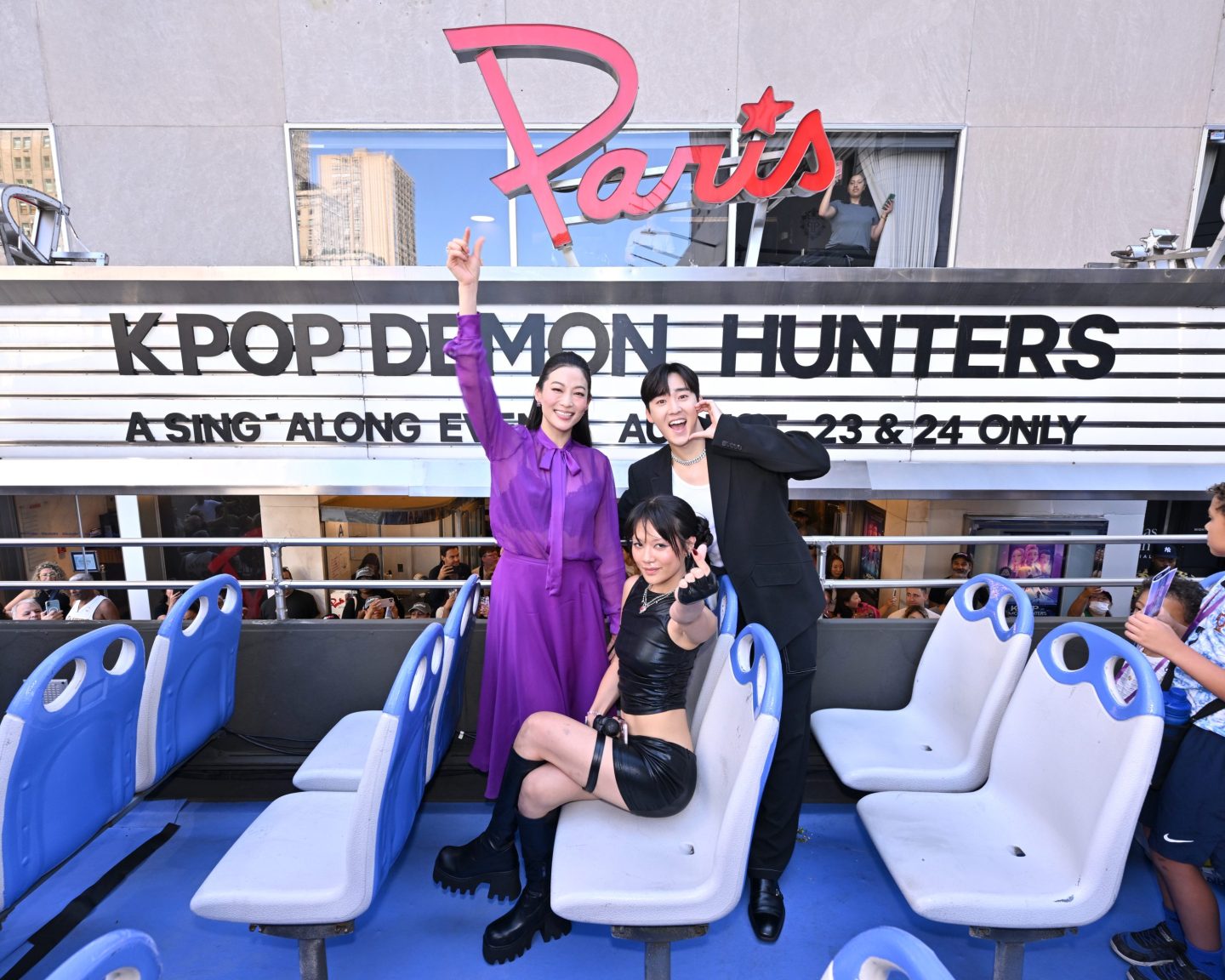 Arden Cho, REI AMI and Kevin Woo raise their hands up on a float in paris in front of a sign that says K-Pop demon hunters.