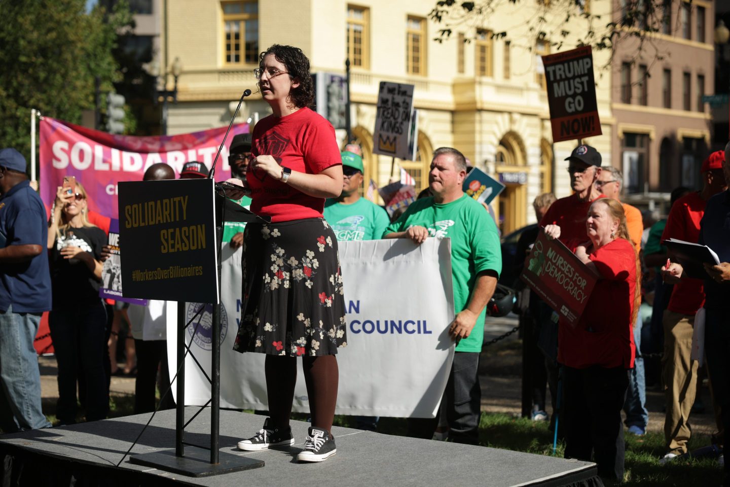 A protestor stands on a stage speaking on a microphone, with protestors behind them holding up a sign that says 'solidarity.'