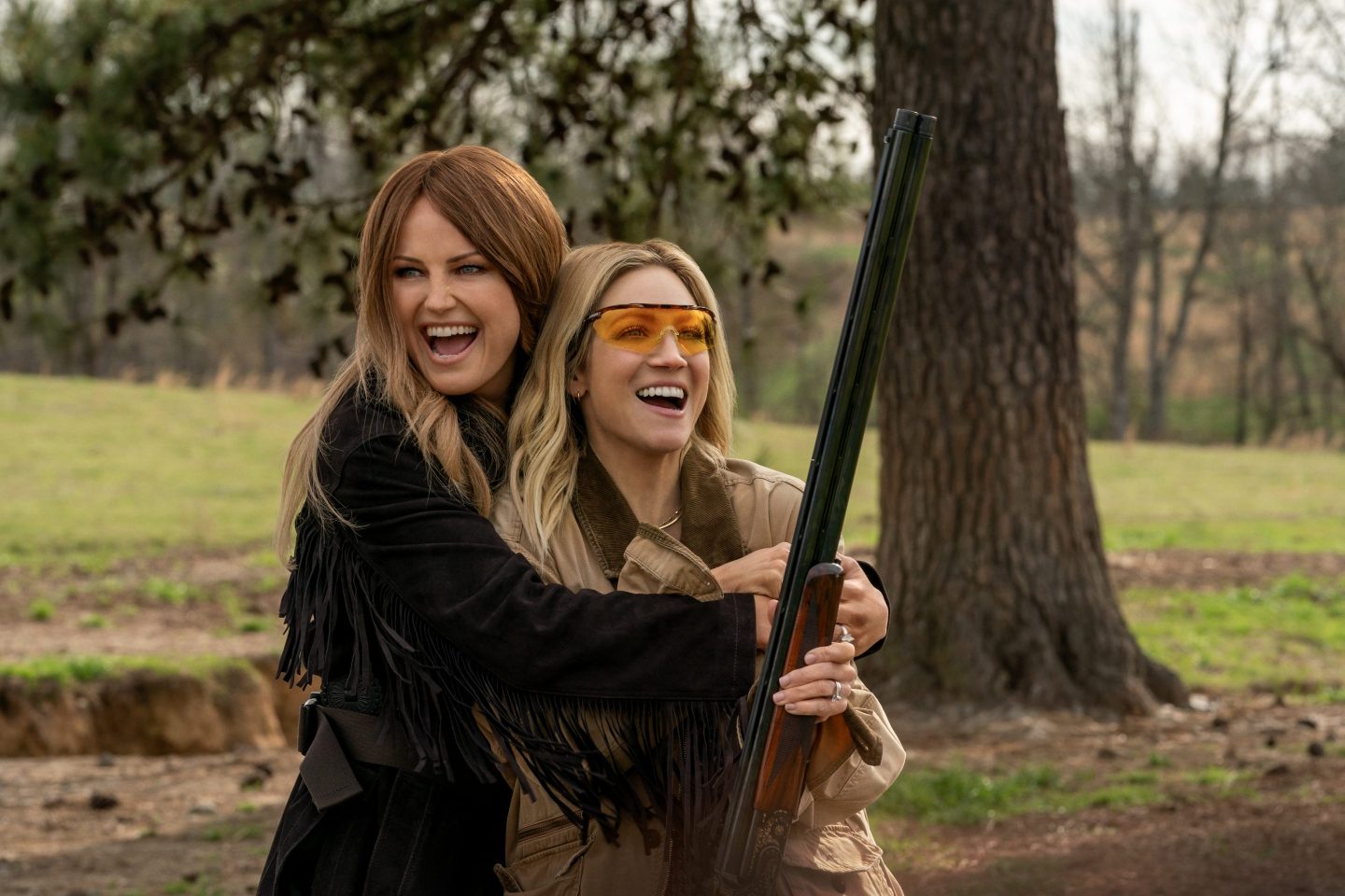Two women embrace and smile while one holds a shotgun