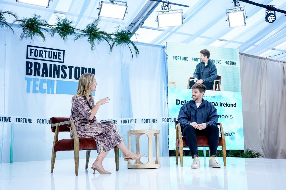 Two people sit facing each other on a stage with a banner reading "Coins2Day Brainstorm Tech" in the background.