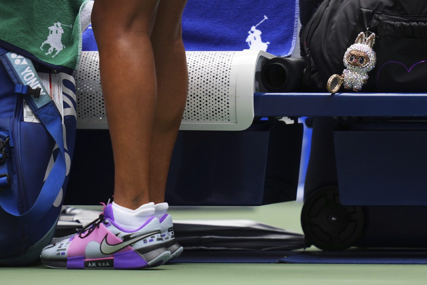 A Labubu doll sits near the legs of Naomi Osaka, of Japan, before her match against Coco Gauff, of the United States, during the U.S> Open. Osaka is wearing purple sparkly shoes.