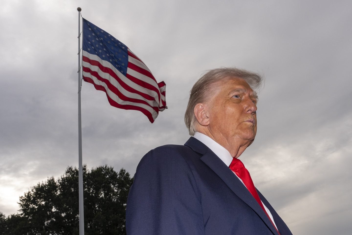 President Donald Trump from a downward angle stands with a straight face next to the American flag.