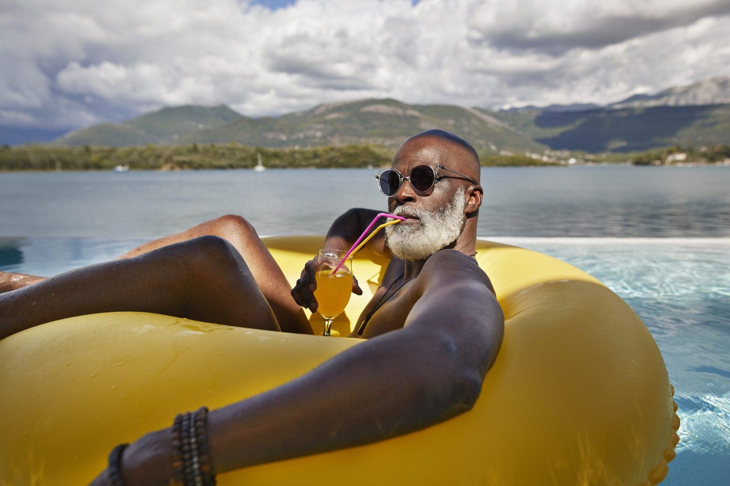 A bald black man with a beautiful white beard wearing sunglasses floats on an inner tube while drinking a fruity beverage