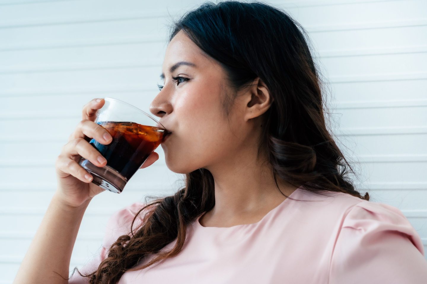 Asian woman drinking soft drink in living room, wearing a pink shirt.