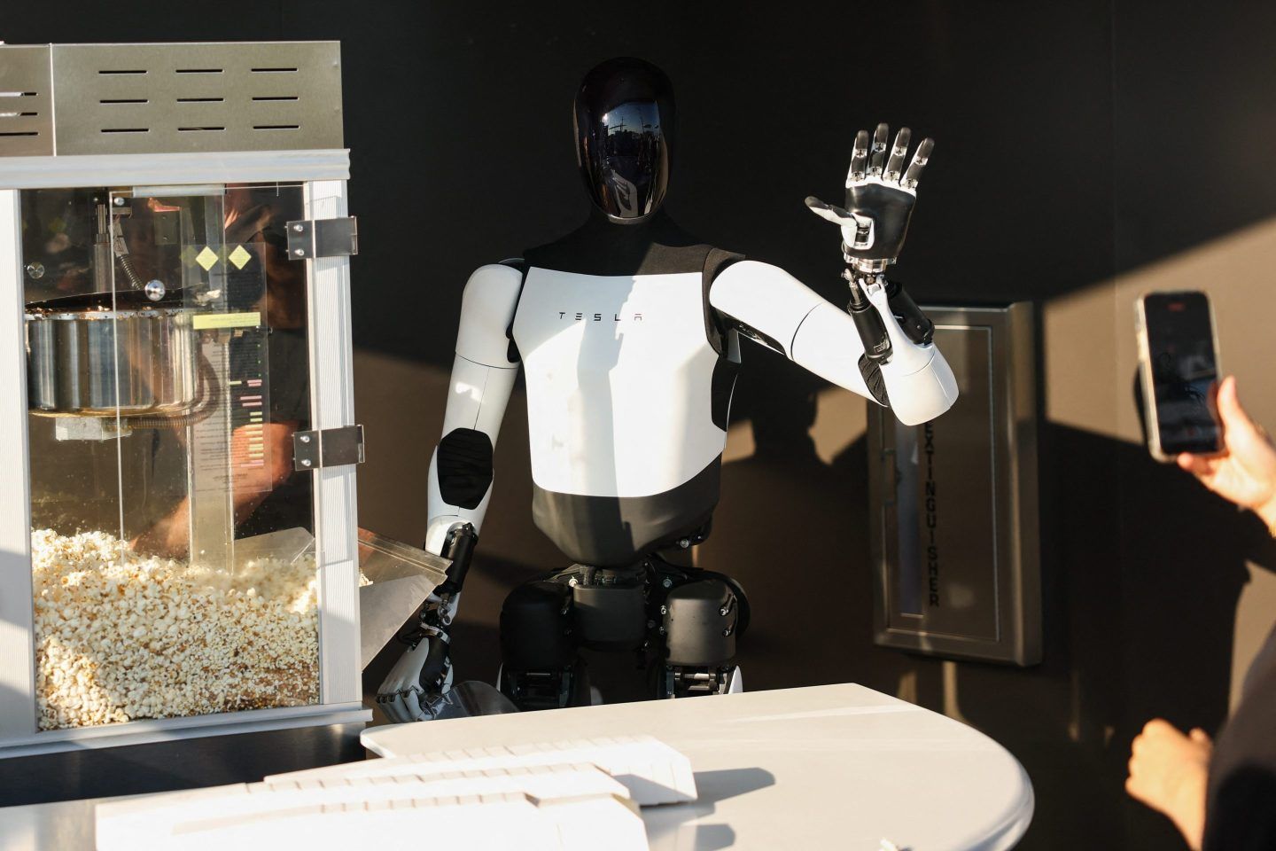 A black-and-white Tesla Optimus bot stands next to a popcorn maker behind a counter and waves.