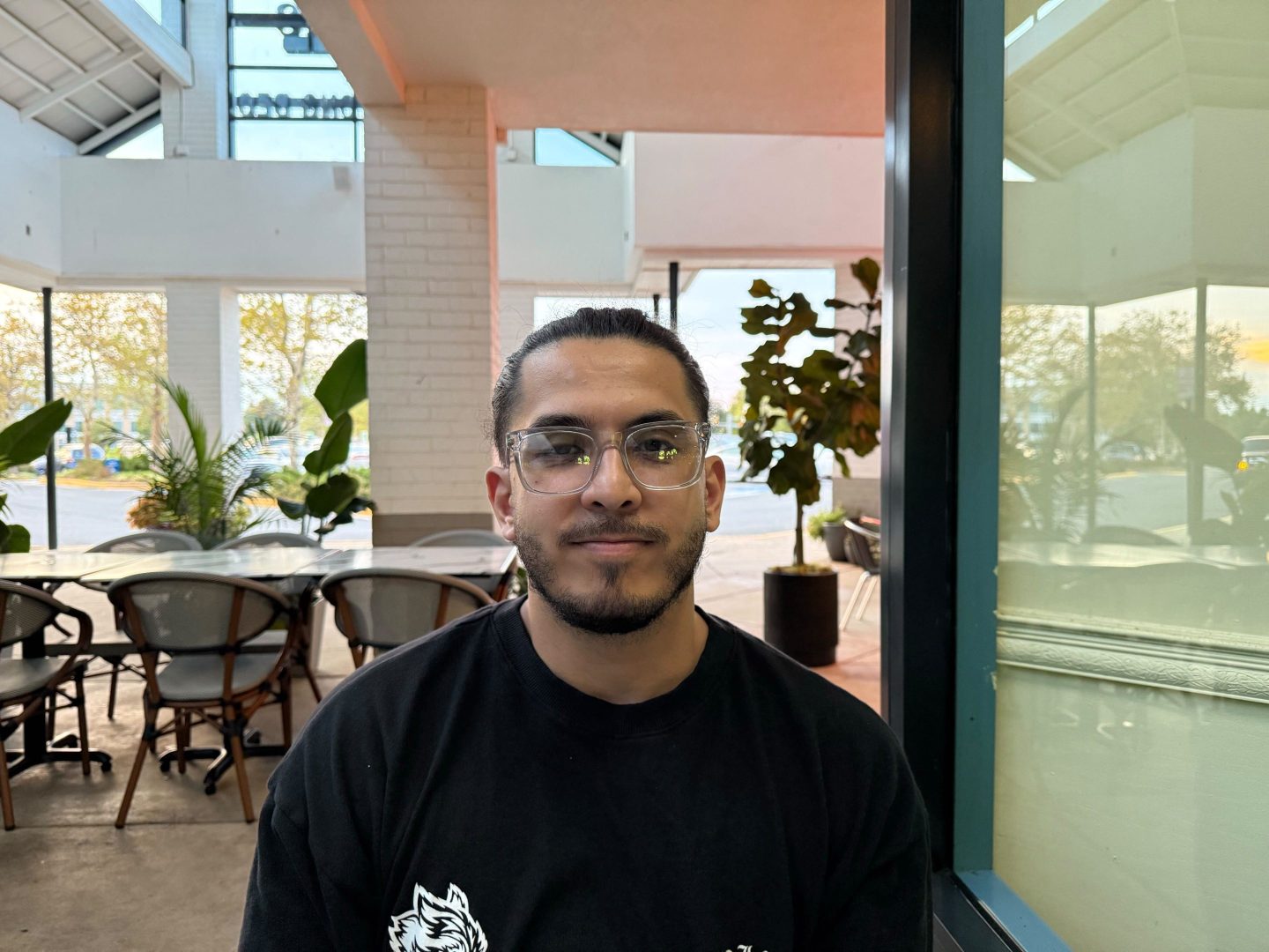 Ali Nasrati sitting next to a reflective glass pane window, in glasses and a black shirt.