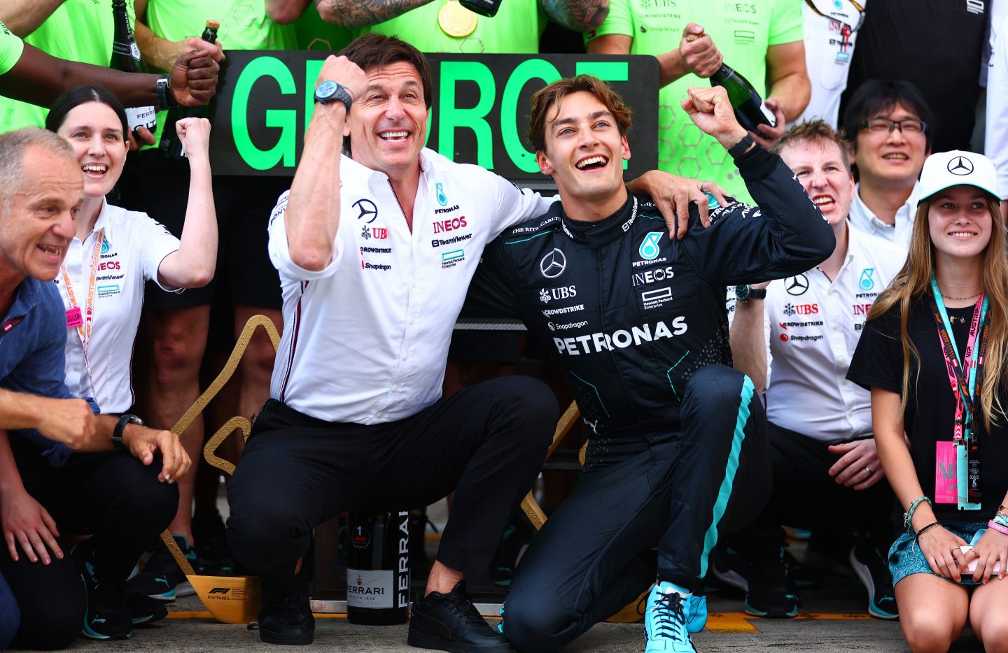 Toto Wolff and George Russell squat and pump their fists into the air, their arms around each other.