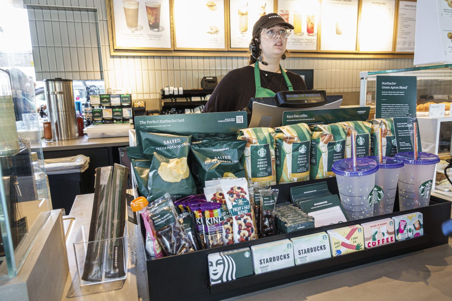 A Starbucks barista stands behind the cash register, looking concerned.