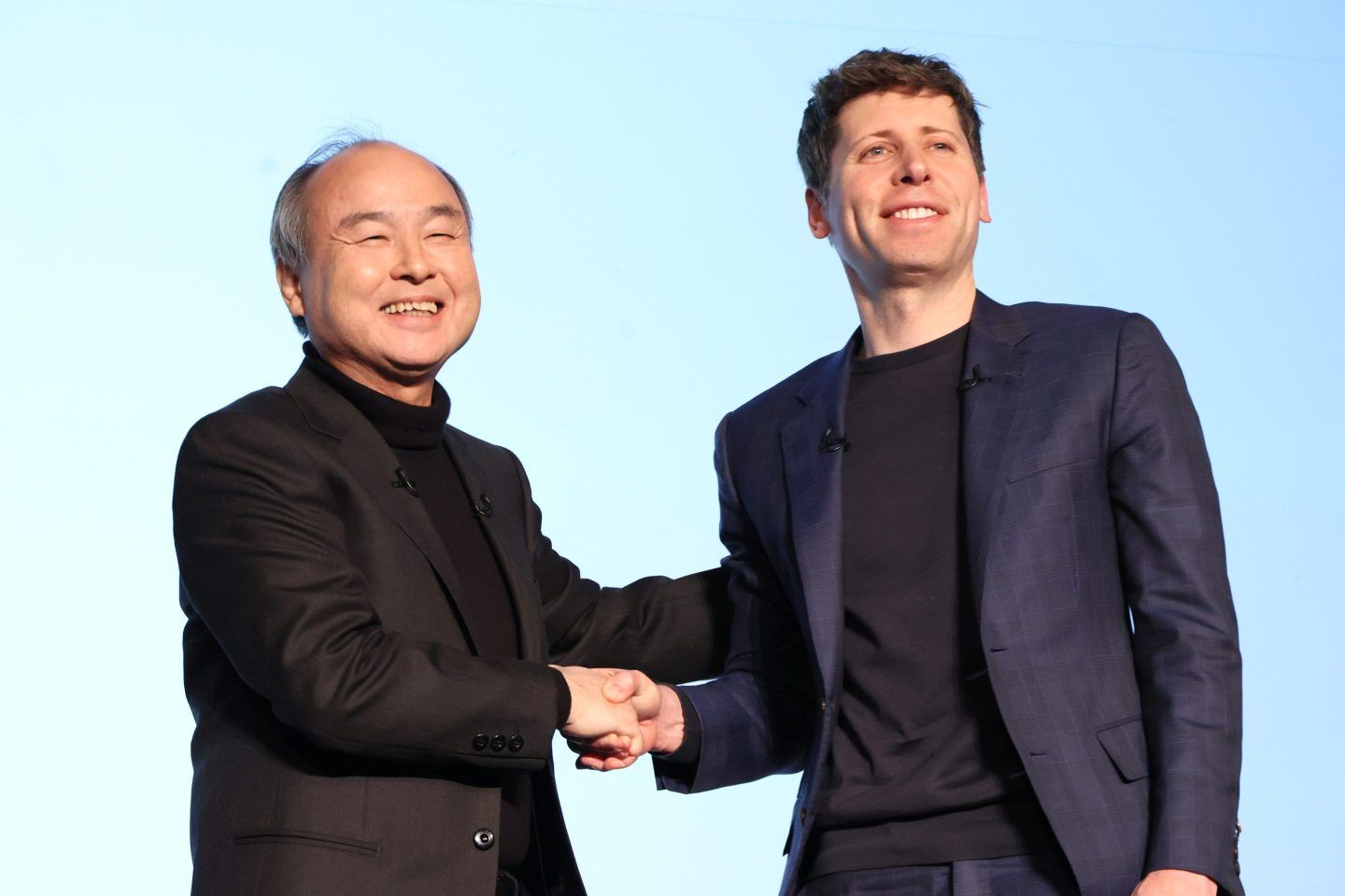 Masayoshi Son and Sam Altman shake hands and smile for the cameras on stage