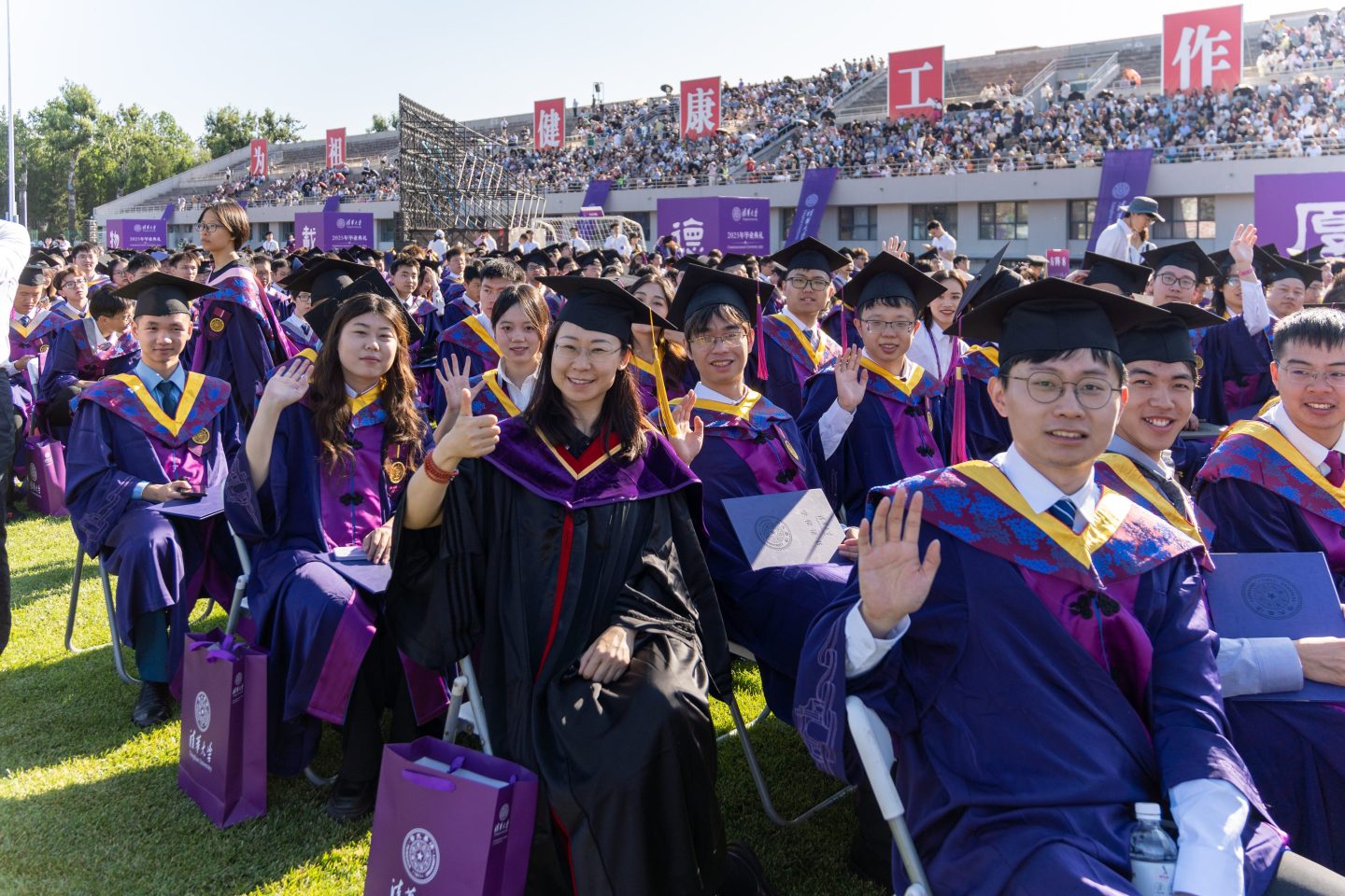 Tsinghua University sit outside in graduation regalia, smiling and waving