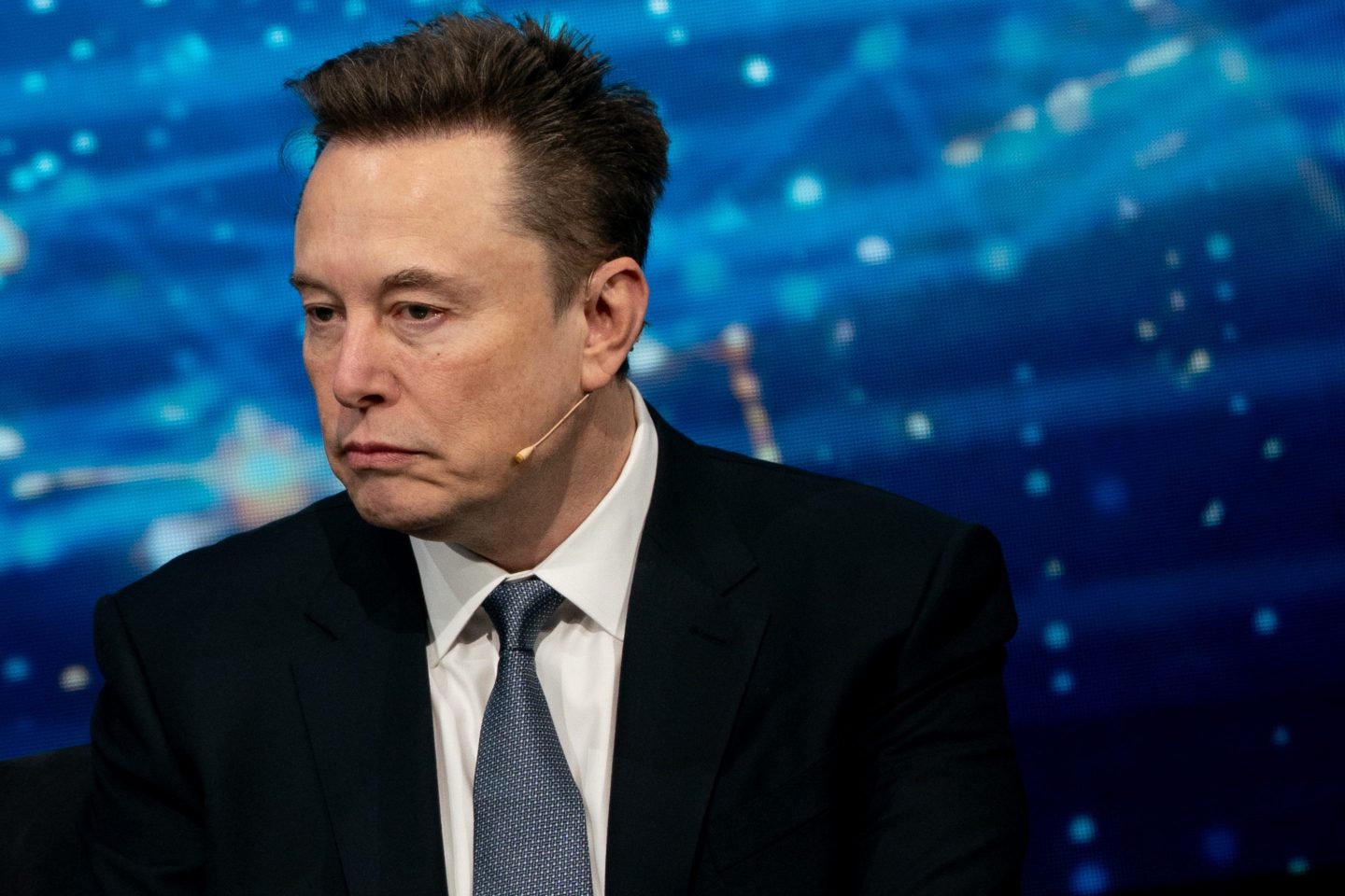 Elon Musk, wearing a suit, looks to the side and frowns.