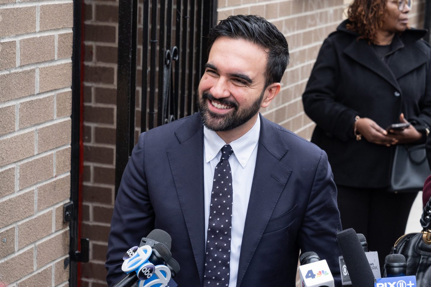 Zohran Mamdani, in front of a brick building, smiles as he holds a press conference.