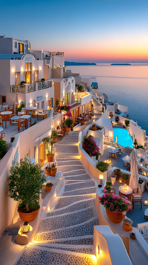 A cobblestone stairway lit by lanterns between whitewashed Santorini buildings overlooking the Aegean Sea at sunset.