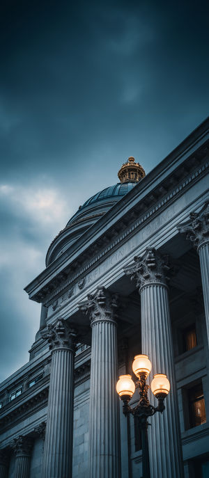 Low-angle view of a historic capitol buildingβs dome and Corinthian columns lit by a vintage street lamp at dusk