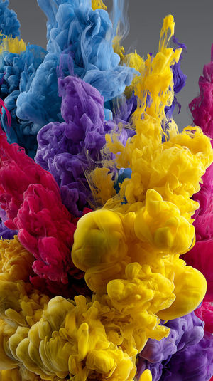 Close-up view of vibrant yellow, purple, blue and red ink clouds swirling in clear water against a neutral background.