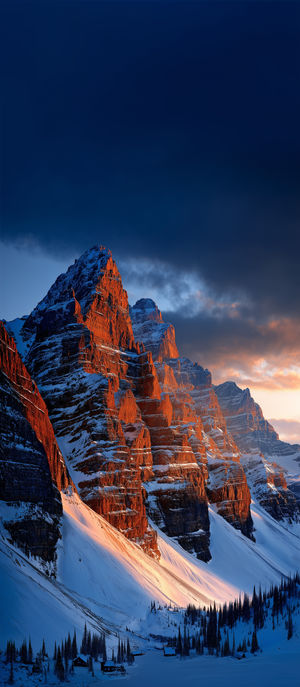 Snow-covered mountain peaks bathed in warm sunrise glow, surrounded by a pine forest and cabins under a deep blue sky.