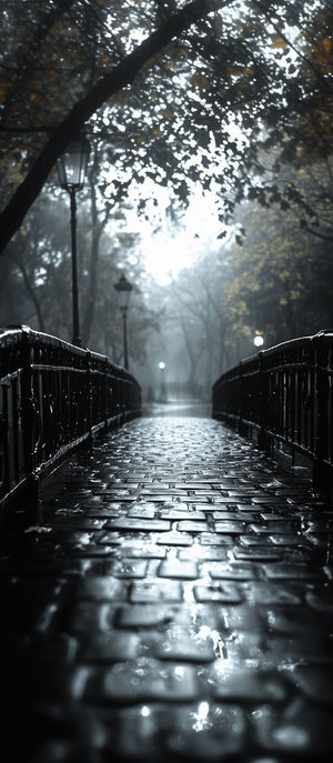 A wet cobblestone bridge in a foggy park at night with glowing lampposts and rain-drenched trees