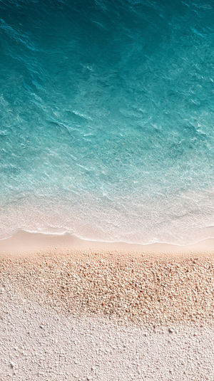 Aerial view of a turquoise sea with crystal-clear waves gently washing over a pale pebble beach under bright sunlight.