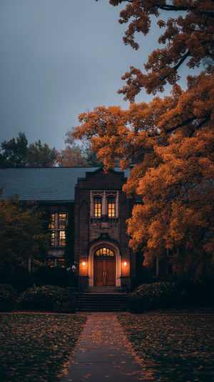 Historic brick campus entrance at dusk with lit wooden doors, orange autumn foliage and glowing lanterns.