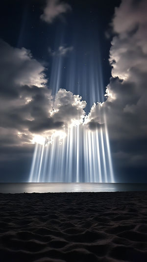 Vertical view of bright celestial light beams streaming through dark clouds onto a calm ocean at night above a sandy shore