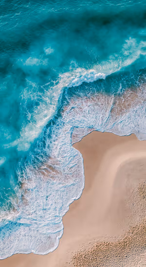 Aerial ocean waves meeting a sandy beach in turquoise and white coastal tones.
