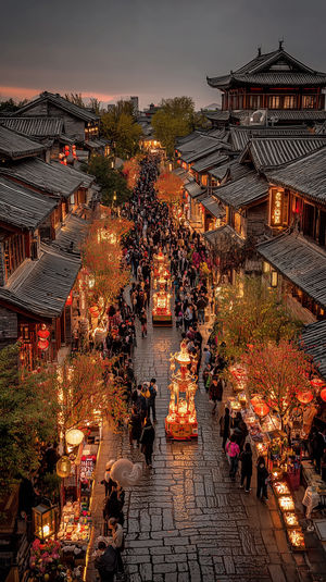 Aerial view of a crowded Chinese night market at dusk with glowing lanterns, festival floats, and tiled roofs.