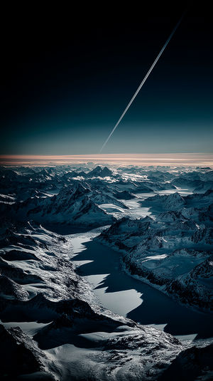 Aerial view of a glacial mountain range with frozen valleys under a twilight teal sky and jet contrail