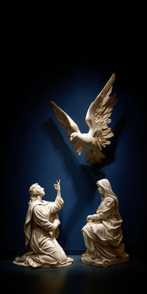 White marble Annunciation sculpture showing two kneeling figures and a hovering dove against a dark blue background.
