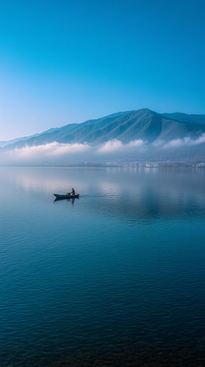 A lone rowboat glides across a calm mountain lake under a clear blue sky, with mist drifting above water.