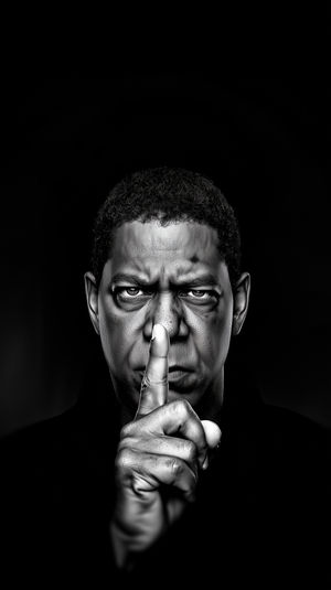 Dramatic black and white close-up of a man holding a finger to his lips against a dark background.