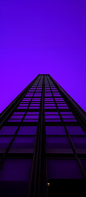 Extreme low-angle view of a modern glass skyscraper with purple-tinted windows soaring into a vibrant violet sky.