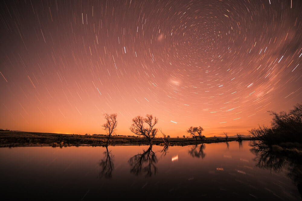 Silhouette of trees reflecting on a lake with stars in the background