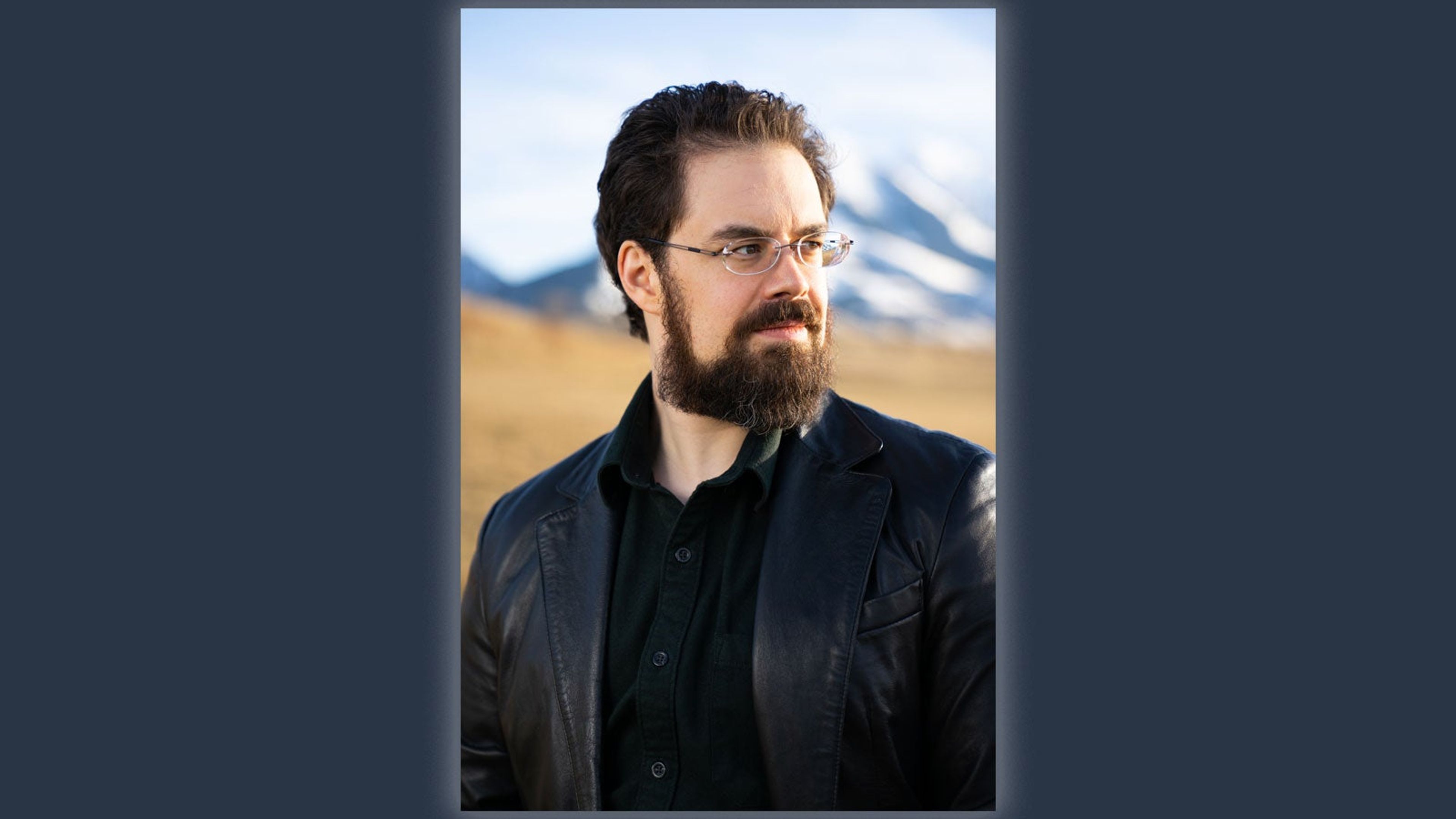 Photograph of Christopher Paolini in a leather jacket and black shirt standing in front of a hill and mountains.