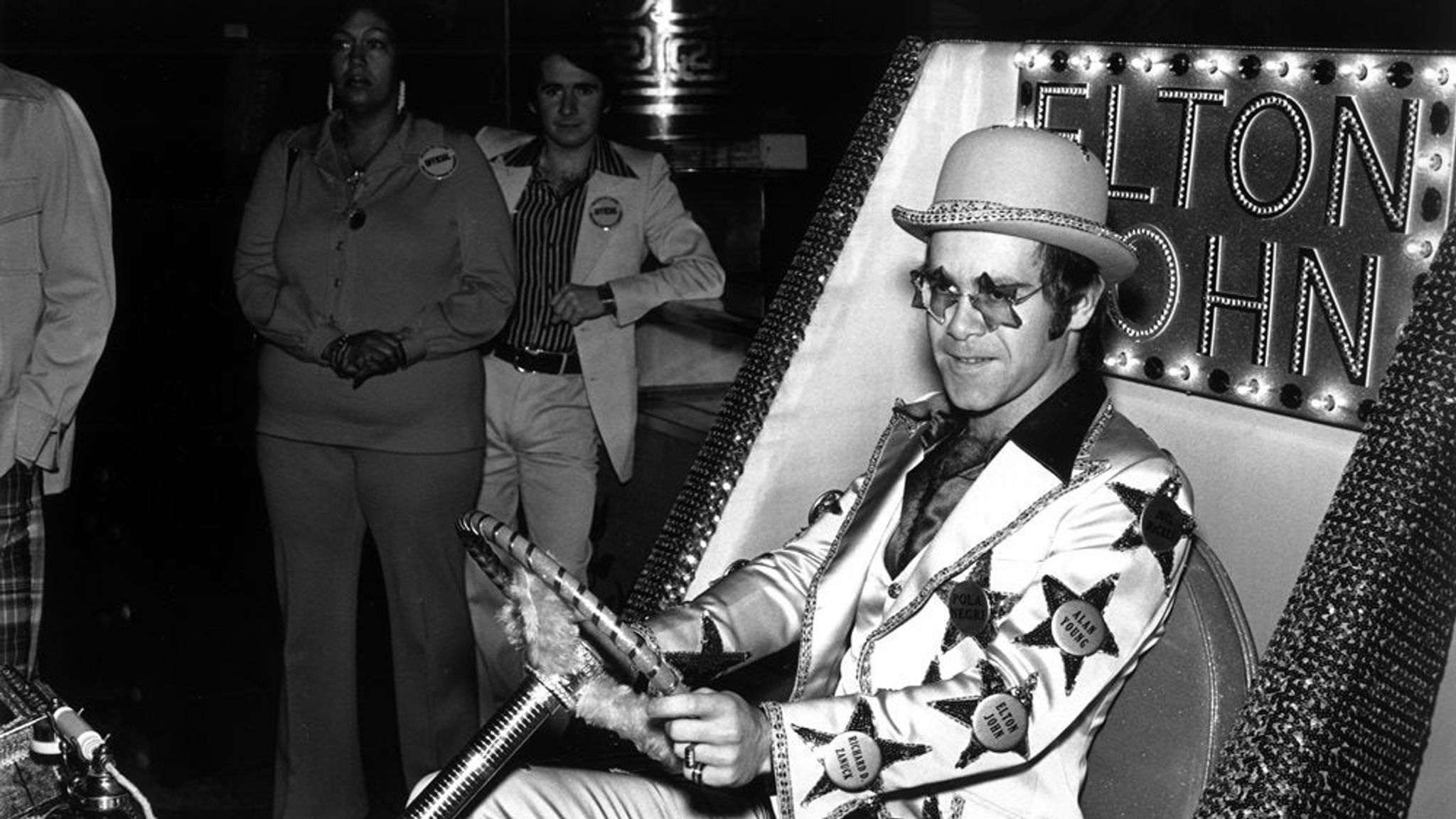 Elton John sitting in a go cart, with his name in lights behind him.